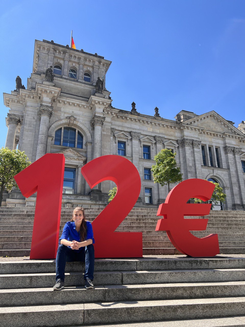 Maja Wallstein MdB auf der Treppe vor einem großen roten 12 Euro Symbol auf der Treppe dem Reichstag