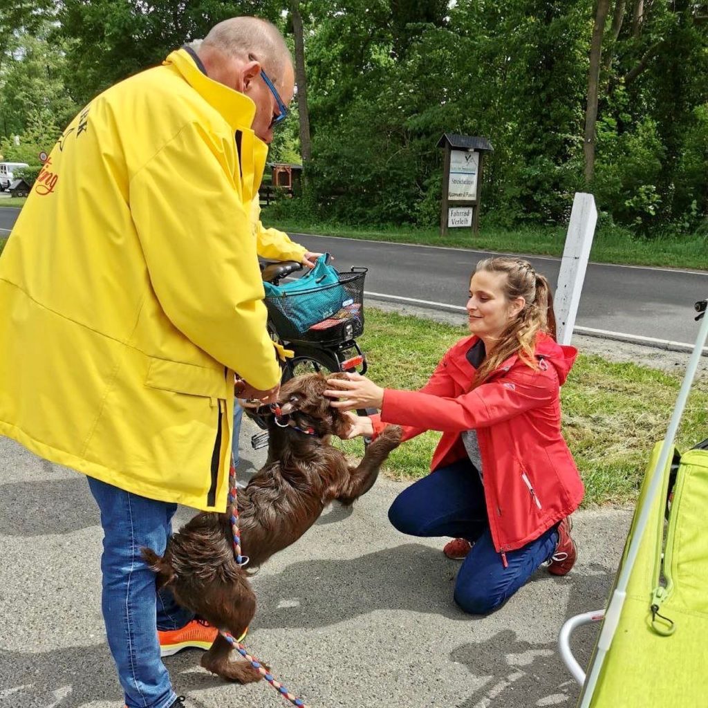 Frau Wallstein wird von einem Hund begrüßt. Zu sehen ist ebenfalls der Hundebesitzer in einer gelben Jacke.