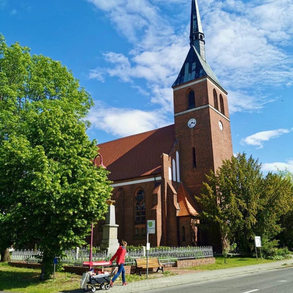 Frau Wallstein zieht ihren Bollerwagen durch eine Ortschaft. Im Hintergrund erstreckt sich eine Kirche.