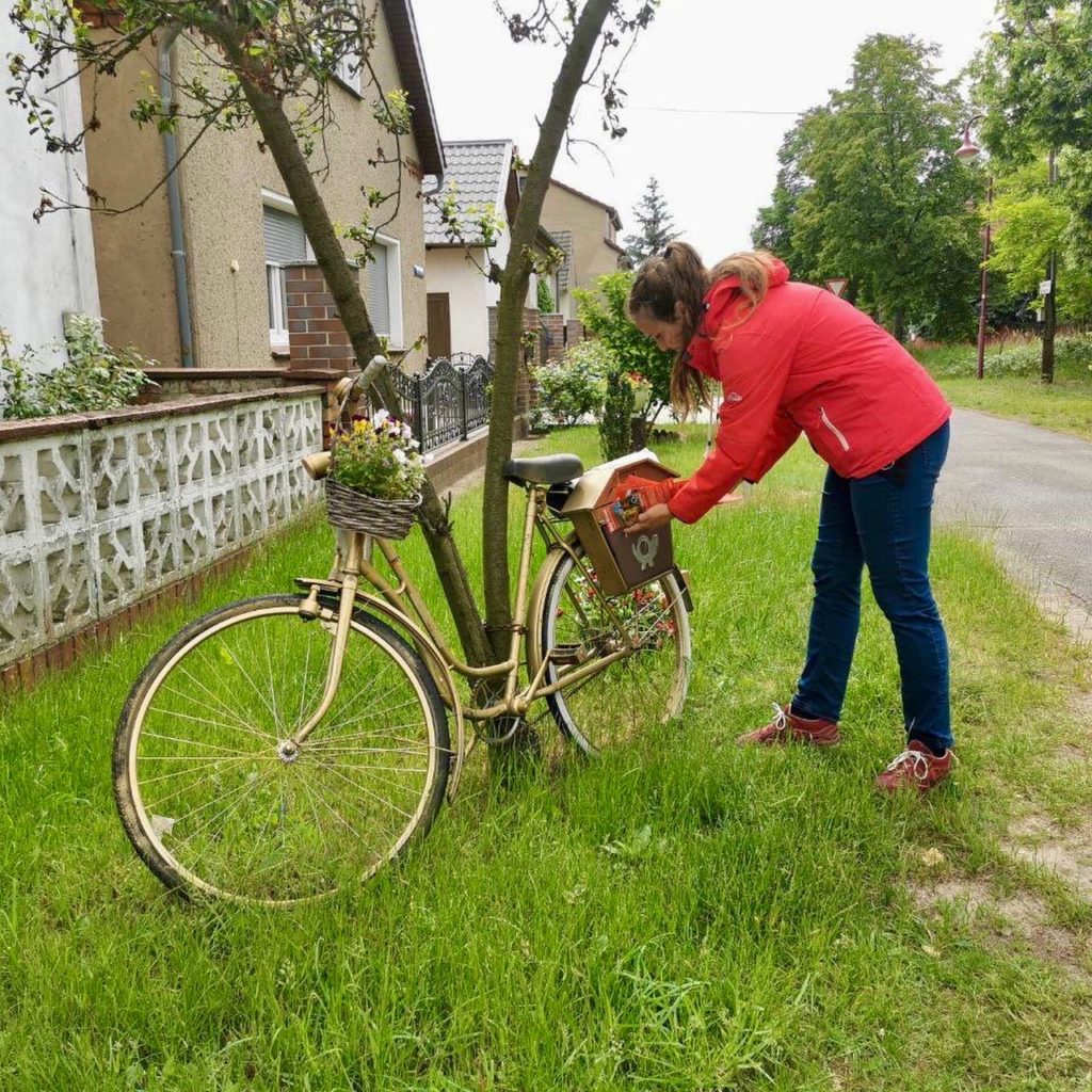 Frau Wallstein wirft einen Flyer in einen Briefkasten ein, welcher an einem Goldenen Fahrrad befestigt ist.