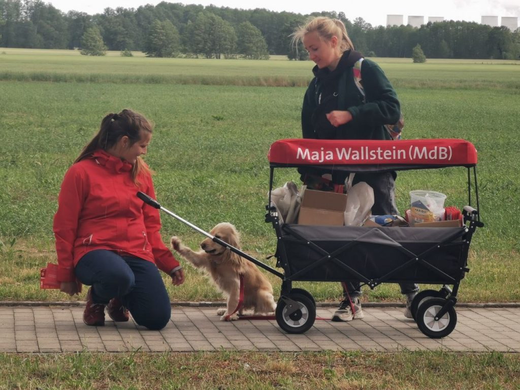 Cocker Spaniel Shelly gibt Maja Wallstein MdB Pfötchen, daneben steht Olya hinter einem Bollerwagen auf einem Fußweg