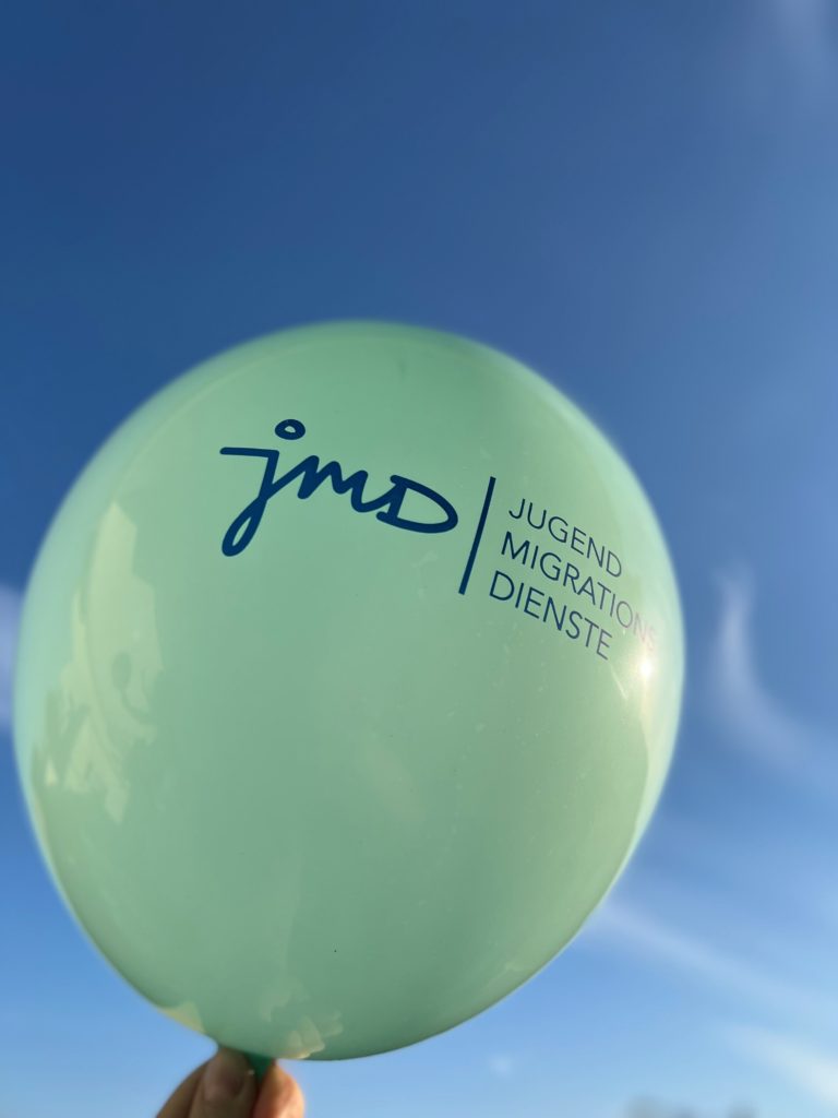 Hand hält grünen Luftballon mit blauem Aufdruck "JMD - Jugendmigrationsdienste" in den blauen Himmel